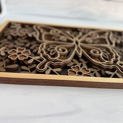 A close-up of a laser-cut wooden decorative panel featuring a butterfly and floral motifs, with layered wood pieces and a light natural wood frame on a white surface.