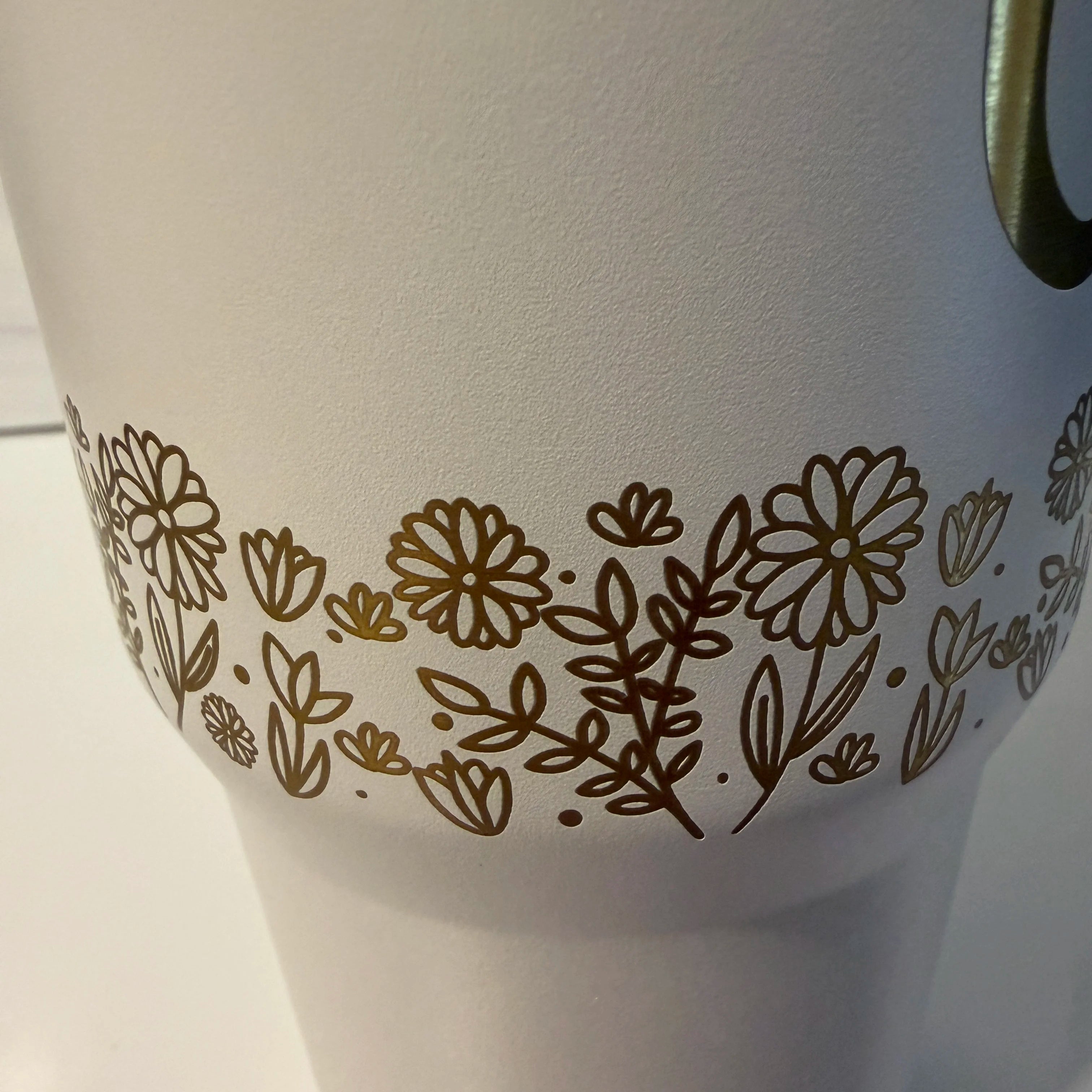 Closeup detail of laser-engraved floral meadow band on white and gold electroplated tumbler showing daisy and wildflower pattern by Cactus Air Designs