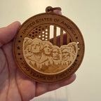 Hand holding USA 250th Anniversary Mount Rushmore wood ornament showing actual size approximately 3.5 inches handcrafted by Cactus Air Designs Arizona
