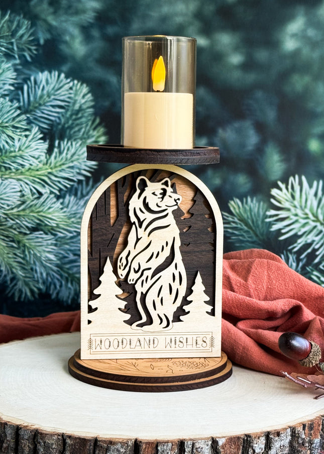 Wooden candle holder with bear design and 'Woodland Wishes' text, surrounded by pine branches and a red cloth.