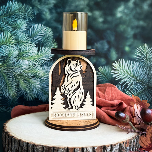 Wooden candle holder with bear design and 'Woodland Wishes' text, surrounded by pine branches and a red cloth.