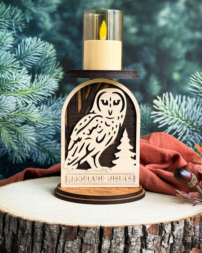 Decorative candle holder with owl design on a wooden base, surrounded by pine branches and leaves.