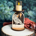 Wooden candle holder with raven design and lit candle against a green foliage background