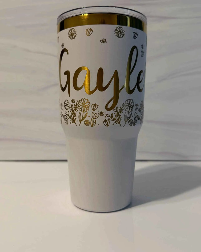 A white insulated tumbler with a clear lid, featuring the name 'Gayle' in gold script, gold floral line art, and a gold rim, set against a light gray wood background.