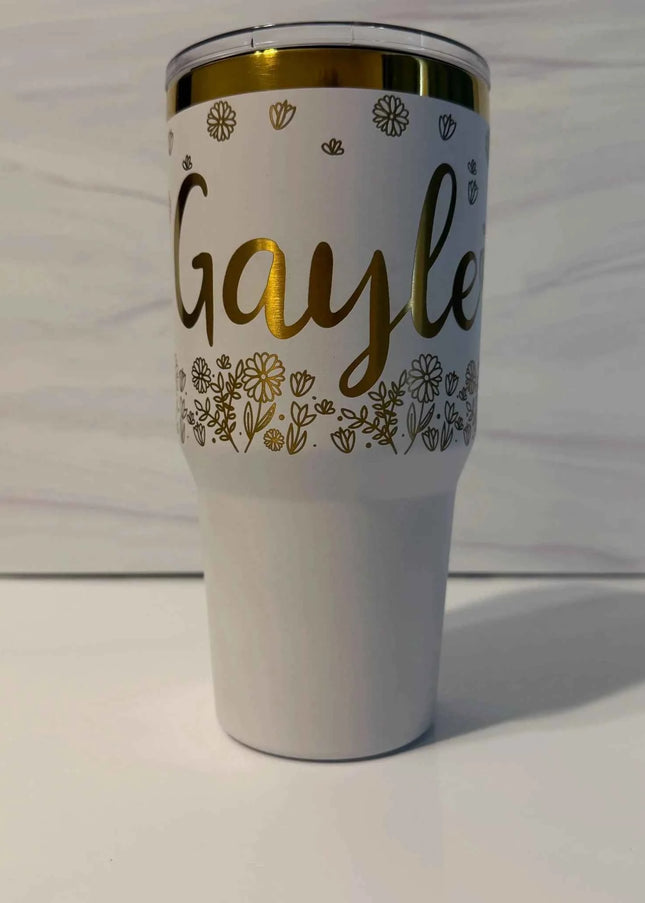 A white insulated tumbler with a clear lid, featuring the name 'Gayle' in gold script, gold floral line art, and a gold rim, set against a light gray wood background.
