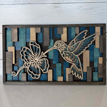 Hummingbird and flower layered wood wall art in cool blue colorway, front view on flat surface showing mosaic stained background and natural maple cutwork
