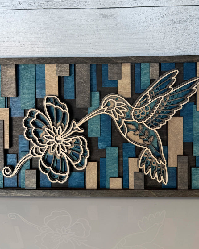 Hummingbird and flower layered wood wall art in cool blue colorway, front view on flat surface showing mosaic stained background and natural maple cutwork
