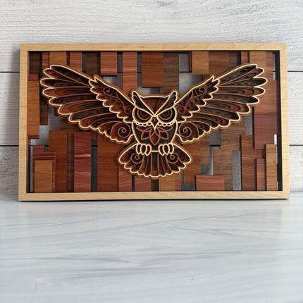 A rectangular wooden wall art piece featuring a laser-cut flying owl design layered over a geometric wood block background, displayed on a light gray surface against a white wood-plank wall.