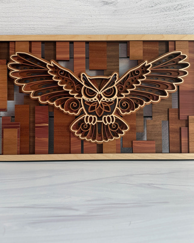 A rectangular wooden wall art piece featuring a laser-cut flying owl design layered over a geometric wood block background, displayed on a light gray surface against a white wood-plank wall.
