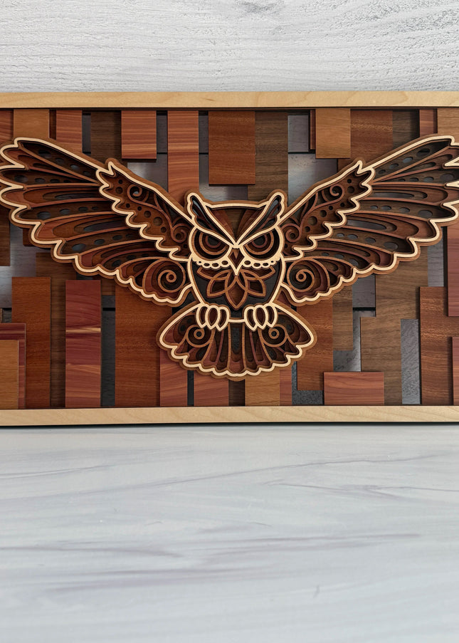A rectangular wooden wall art piece featuring a laser-cut flying owl design layered over a geometric wood block background, displayed on a light gray surface against a white wood-plank wall.