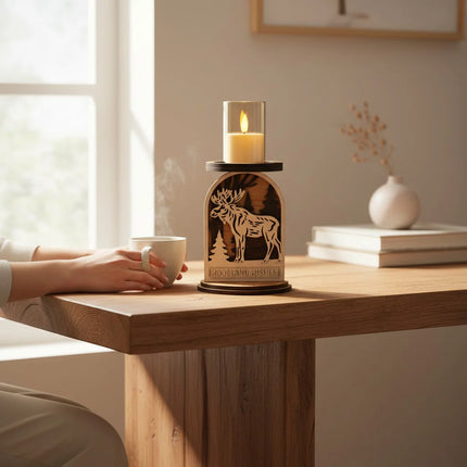 A wooden moose-shaped candle holder with a LED candle in a glass holder on top, placed on a wooden table in a warm, sunlit room. A person's hands and arm are visible holding a white mug at the table, with books and a small vase in the background.