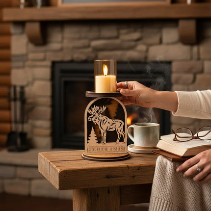A wooden moose-shaped candle holder with a glass LED candle on top, placed on a rustic wooden table in front of a cozy stone fireplace. A person's hand is adjusting the candle, with a mug, book, and glasses nearby.