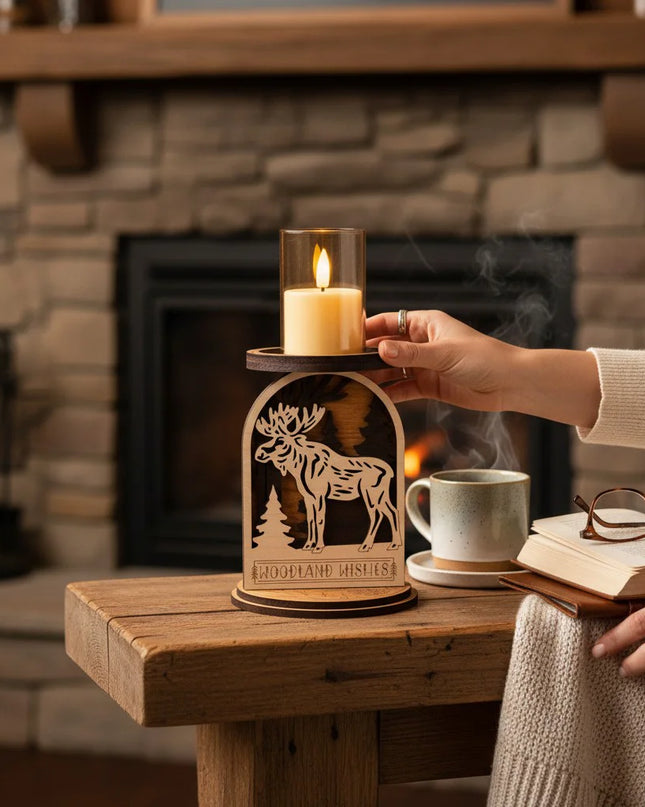 A wooden moose-shaped candle holder with a glass LED candle on top, placed on a rustic wooden table in front of a cozy stone fireplace. A person's hand is adjusting the candle, with a mug, book, and glasses nearby.