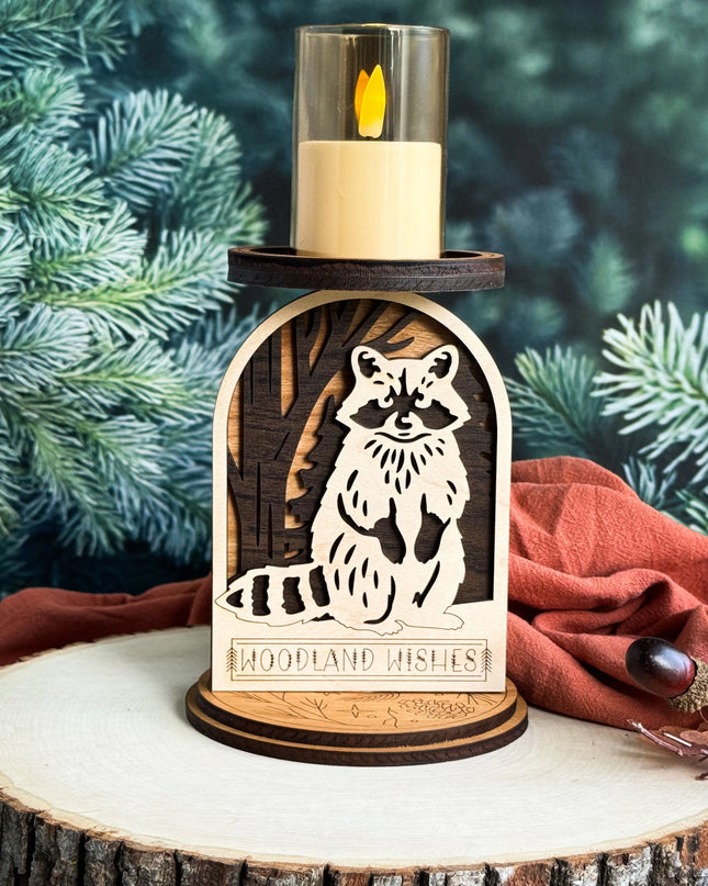 A wooden candle holder with a laser-cut raccoon design and the words 'WOODLAND' on the front, holding a lit cream candle in a clear glass holder. The holder is displayed on a rustic wood slice with evergreen branches and a rust-colored fabric in the background.