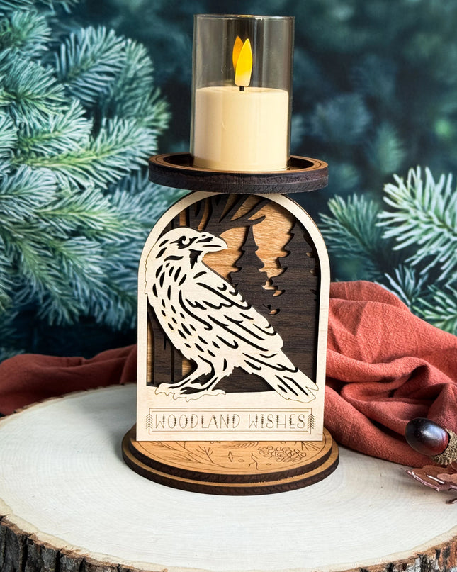 A wooden raven candle holder with a lit cream candle in a glass holder, styled on a rustic wood slice with a rust-colored cloth and evergreen branches in the background.