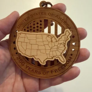 Round layered wooden ornament held in an adult hand for scale, featuring a raised map of the contiguous United States with engraved state borders over a cut-through American flag. The border reads “United States of America” and “250 Years of Freedom.”