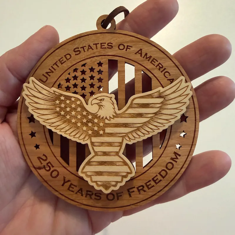 Round layered wooden ornament held in an adult hand for scale, featuring a raised bald eagle patterned with stars and stripes over a cut-through American flag. The border reads “United States of America” and “250 Years of Freedom.”