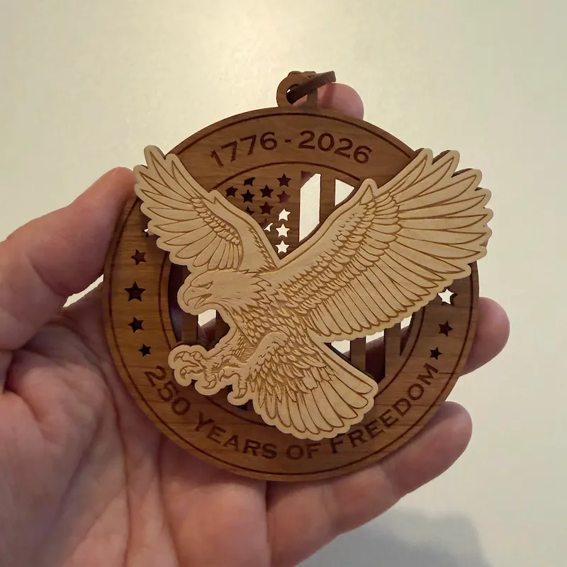 Round layered wooden ornament held in an adult hand for scale, featuring a raised bald eagle swooping forward over a cut-through American flag. The border reads “1776–2026” and “250 Years of Freedom.”
