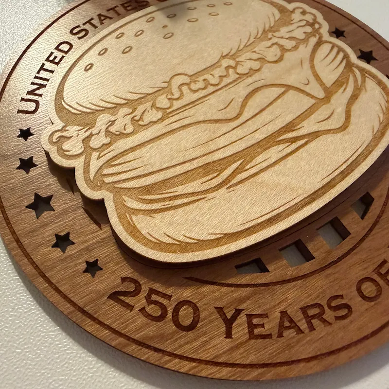 Close-up detail of a layered wooden ornament showing a raised cheeseburger with a laser-engraved sesame seed bun and stacked layers of lettuce, cheese, toppings, and patties over a darker wood base.
