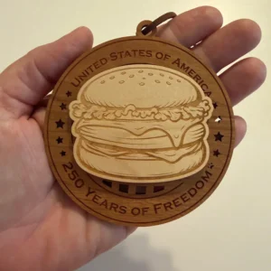Round layered wooden ornament held in an adult hand for scale, featuring a raised, laser-engraved cheeseburger surrounded by star cutouts. The border reads “United States of America” and “250 Years of Freedom.”