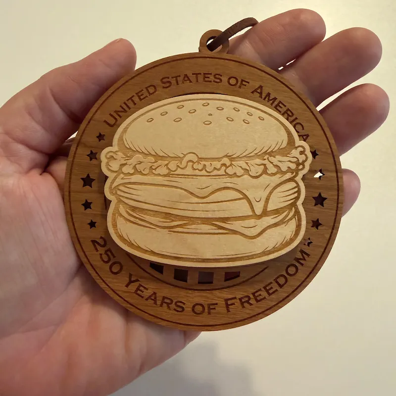 Round layered wooden ornament held in an adult hand for scale, featuring a raised, laser-engraved cheeseburger surrounded by star cutouts. The border reads “United States of America” and “250 Years of Freedom.”