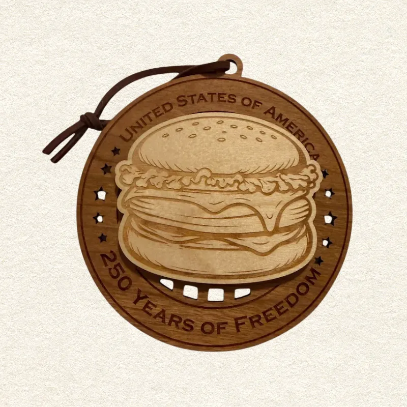 Round two-layer wooden ornament with "United States of America" and "250 Years of Freedom" engraved on the border, featuring a laser-cut flag with cut-through stripes and star field, topped with a detailed cheeseburger, hung on a brown cord
