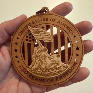 Round layered wooden ornament held in an adult hand for scale, depicting service members raising the American flag at Iwo Jima over a cut-through flag background. The border reads “United States of America” and “250 Years of Freedom.”