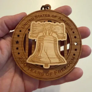 Round layered wooden ornament held in an adult hand for scale, featuring a raised Liberty Bell with its wooden support and iconic crack over a cut-through American flag. The border reads “United States of America” and “250 Years of Freedom.”