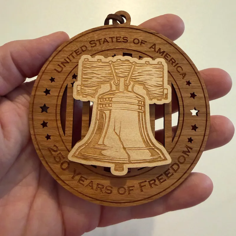 Round layered wooden ornament held in an adult hand for scale, featuring a raised Liberty Bell with its wooden support and iconic crack over a cut-through American flag. The border reads “United States of America” and “250 Years of Freedom.”