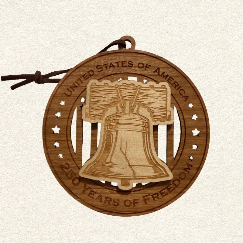Round two-layer wooden ornament with "United States of America" and "250 Years of Freedom" engraved on the border, featuring a laser-cut flag with cut-through stripes and star field, topped with a detailed Liberty Bell and its crack on it, hung on a brown leather cord