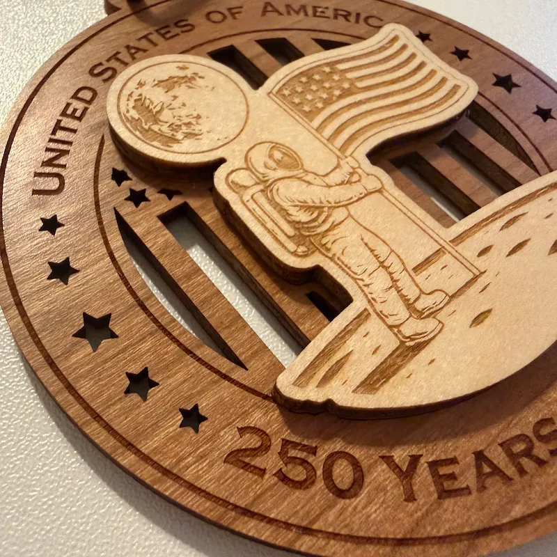 Close-up detail of a layered wooden ornament showing a raised astronaut standing on the moon and planting the American flag, with laser-engraved spacesuit, lunar surface, and Earth details over a cut-through flag background.