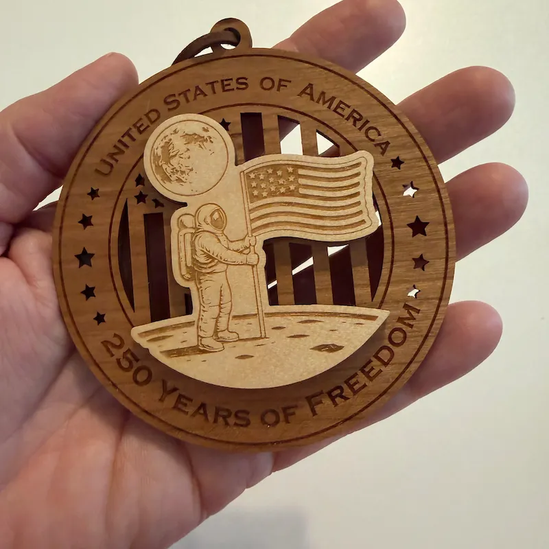 Round layered wooden ornament held in an adult hand for scale, showing an astronaut planting the American flag on the moon with Earth overhead. The border reads “United States of America” and “250 Years of Freedom.”
