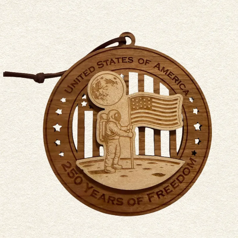 Round two-layer wooden ornament with "United States of America" and "250 Years of Freedom" engraved on the border, featuring a laser-cut flag with cut-through stripes and star field, topped with a detailed of the moon landing and planting the USA flag, hung on a brown cord
