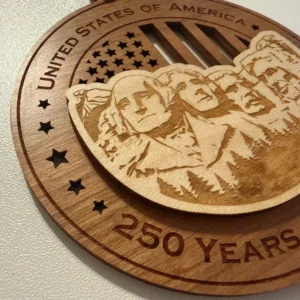 Close-up detail of a layered wooden ornament showing a raised Mount Rushmore with laser-engraved portraits of George Washington, Thomas Jefferson, Theodore Roosevelt, and Abraham Lincoln over a cut-through American flag background.