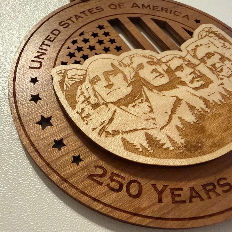 Close-up detail of a layered wooden ornament showing a raised Mount Rushmore with laser-engraved portraits of George Washington, Thomas Jefferson, Theodore Roosevelt, and Abraham Lincoln over a cut-through American flag background.