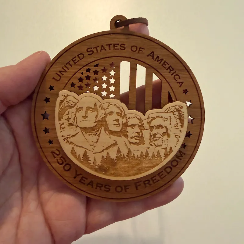 Round layered wooden ornament held in an adult hand for scale, featuring Mount Rushmore with the four presidents and pine trees over a cut-through American flag. The border reads “United States of America” and “250 Years of Freedom.”