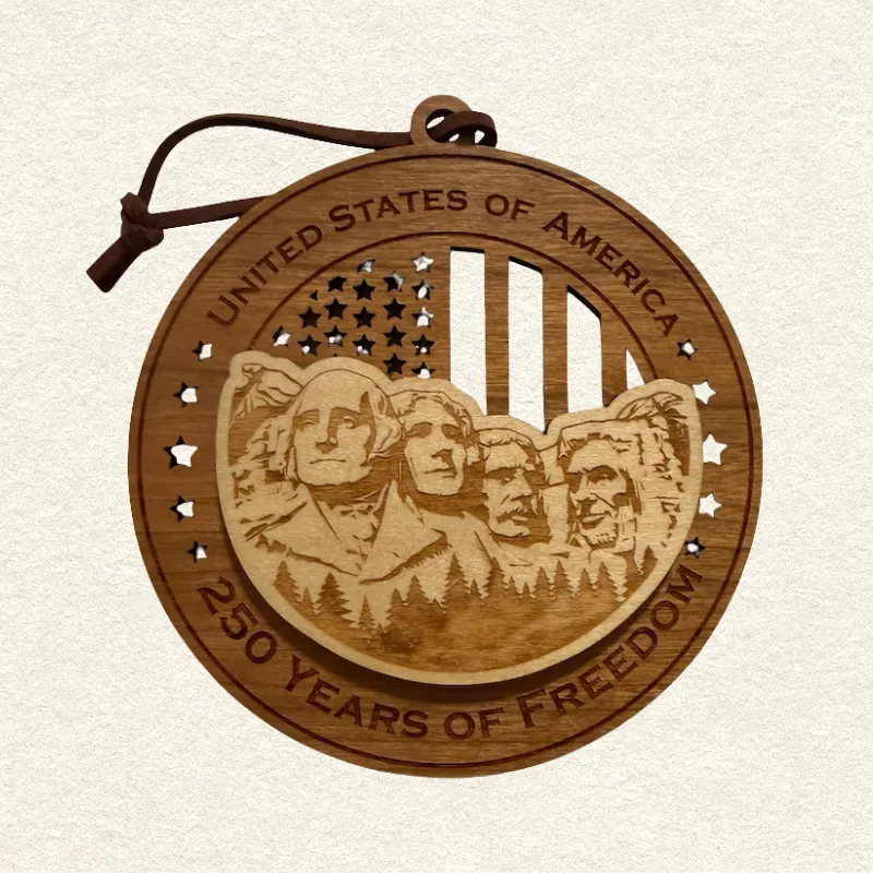 Round two-layer wooden ornament with "United States of America" and "250 Years of Freedom" engraved on the border, featuring a laser-cut flag with cut-through stripes and star field, topped with a detailed Mount Rushmore with the presidents George Washington, Thomas Jefferson, ,Theodore Roosevelt and Abraham Lincoln , hung on a brown leather cord