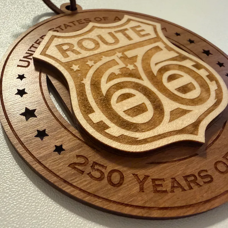 Close-up detail of a layered wooden ornament showing a raised Route 66 highway shield with large laser-engraved numerals, lettering, stars, and shaded accents over a darker wood base with star cutouts.