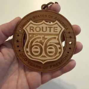 Round layered wooden ornament held in an adult hand for scale, featuring a raised Route 66 highway shield surrounded by star cutouts. The border reads “United States of America” and “250 Years of Freedom.”