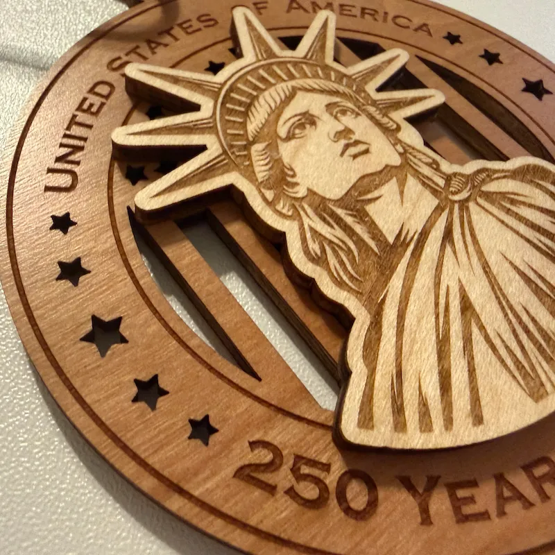 Close-up detail of a layered wooden ornament showing a raised Statue of Liberty with finely laser-engraved facial features, crown, hair, and flowing robe over a cut-through American flag background.