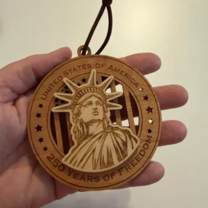 Round layered wooden ornament with a brown hanging loop, held in an adult hand for scale and featuring a raised Statue of Liberty over a cut-through American flag. The border reads “United States of America” and “250 Years of Freedom.”