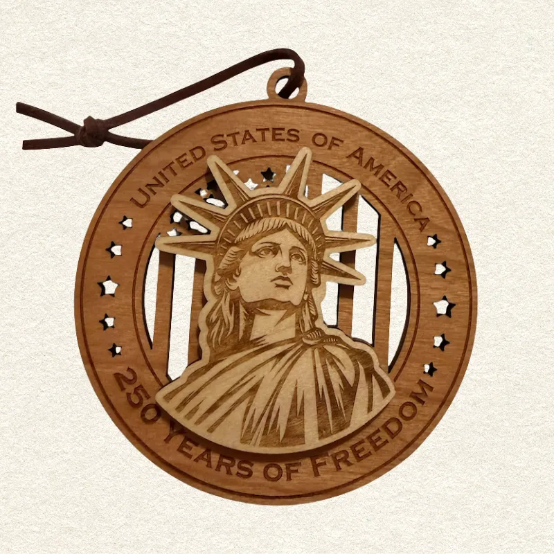 Round two-layer wooden ornament with "United States of America" and "250 Years of Freedom" engraved on the border, featuring a laser-cut flag with cut-through stripes and star field, topped with a detailed Statue of Liberty, hung on a brown cord