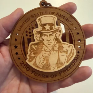 Round layered wooden ornament held in an adult hand for scale, featuring Uncle Sam pointing forward over a cut-through American flag. The border reads “United States of America” and “250 Years of Freedom.”