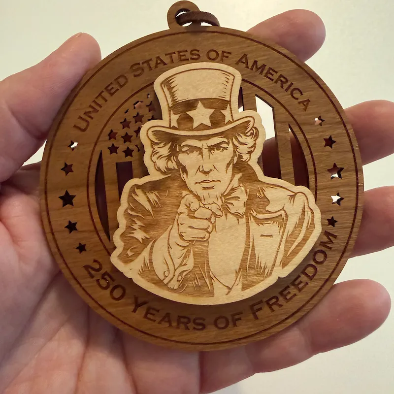 Round layered wooden ornament held in an adult hand for scale, featuring Uncle Sam pointing forward over a cut-through American flag. The border reads “United States of America” and “250 Years of Freedom.”