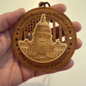 Round layered wooden ornament held in an adult hand for scale, featuring a raised, finely engraved U.S. Capitol over a cut-through American flag. The border reads “United States of America” and “250 Years of Freedom.”