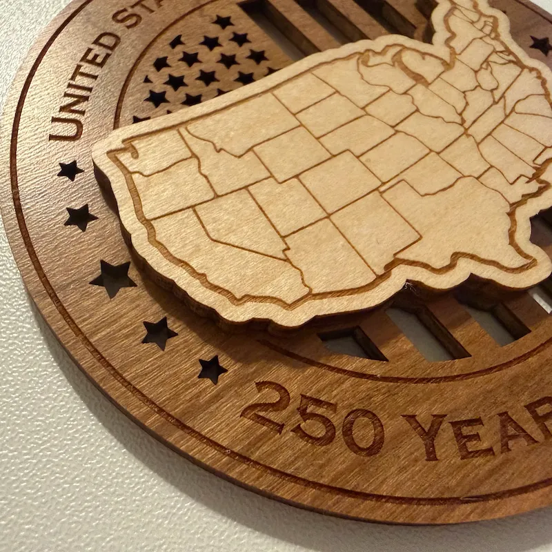 Close-up detail of a layered wooden ornament showing a raised USA map with laser-engraved state lines over cut-through American flag stripes, surrounded by star cutouts and deeply engraved border lettering.