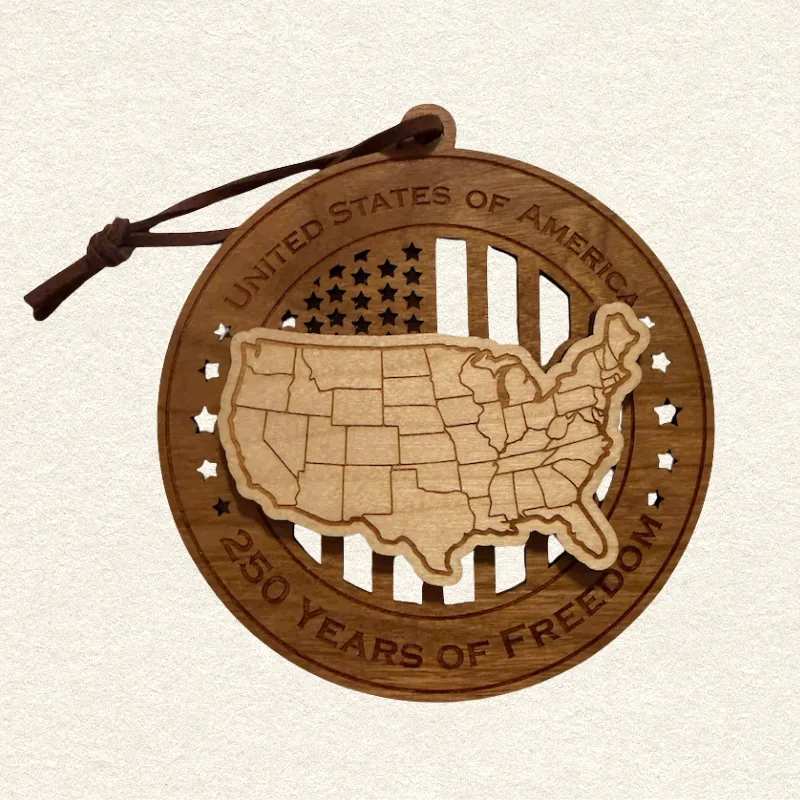 Round two-layer wooden ornament with "United States of America" and "250 Years of Freedom" engraved on the border, featuring a laser-cut flag with cut-through stripes and star field, topped with a detailed USA map showing all state lines, hung on a brown cord